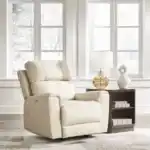 Dahlmoore Power Rocker Recliner in light almond leather, sitting in a bright, modern living room setting with large windows. The chair is slightly reclined. Furniture stores Tampa display.
