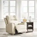 Dahlmoore Power Rocker Recliner in light almond leather, sitting in a bright, modern living room setting with large windows. The chair is slightly reclined. Furniture stores Tampa display.