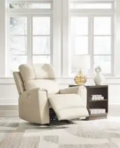 Dahlmoore Power Rocker Recliner in light almond leather, sitting in a bright, modern living room setting with large windows. The chair is slightly reclined. Furniture stores Tampa display.