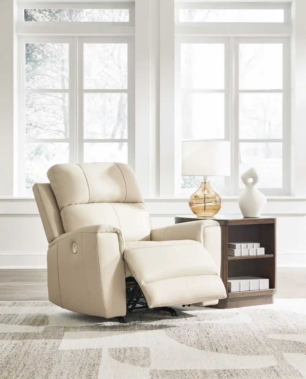 Dahlmoore Power Rocker Recliner in light almond leather, sitting in a bright, modern living room setting with large windows. The chair is slightly reclined. Furniture stores Tampa display.