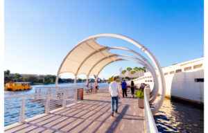 People walking and cycling on the Tampa Riverwalk promenade under a white arch canopy