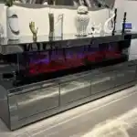 A close-up of a dark gray mirrored TV stand's electric fireplace with a red glow and the remote control on top.