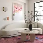 Patrizia 36-inch Faux Travertine Concrete Indoor/Outdoor Round Coffee Table, modern curved sofa, pink floral rug, and contemporary artwork in a luxury living room setting.