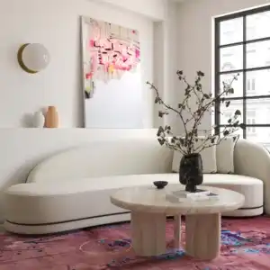 Patrizia 36-inch Faux Travertine Concrete Indoor/Outdoor Round Coffee Table, modern curved sofa, pink floral rug, and contemporary artwork in a luxury living room setting.