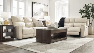 Modern cream-colored power reclining sofa and loveseat set in a bright, sunlit living room with contemporary decor.