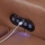 Close-up of the illuminated power control panel (recliner buttons) and integrated USB charging port on the armrest of the Caramel Power Sofa, with a cable connected.
