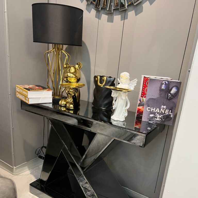 A stylish black console table with a geometric design, decorated with modern home decor items including a black lamp with a gold base, a gold bear figurine, a black and gold vase, a white angel figurine, and two Chanel coffee table books, set against a gray wall with a reflective fragmented mirror.