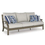 Three-quarter angle view of Milas aluminum outdoor sofa showing wood-look armrest, beige cushion depth, and blue decorative pillows