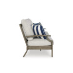 Side profile of Milas beige outdoor couch showing wood-look aluminum armrest, thick beige cushion, and blue striped throw pillow